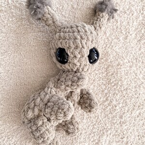 May include: A beige crochet plush toy featuring a textured yarn body, two tall ears topped with fluffy gray pom-poms, and two large, smooth, black plastic eyes. The creature has a rounded head and a soft, thick, looped yarn construction.