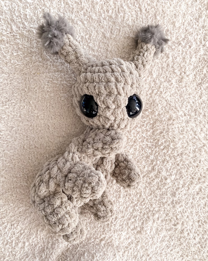 May include: A beige crochet plush toy featuring a textured yarn body, two tall ears topped with fluffy gray pom-poms, and two large, smooth, black plastic eyes. The creature has a rounded head and a soft, thick, looped yarn construction.