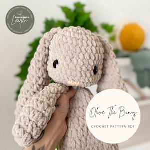 Peut inclure: Un jouet en peluche de lapin crocheté de couleur brun clair. Le lapin a de grandes oreilles tombantes et une expression douce. Le jouet est tenu par une main avec un petit tatouage. L'image comprend le texte "Olive The Bunny CROCHET PATTERN PDF".