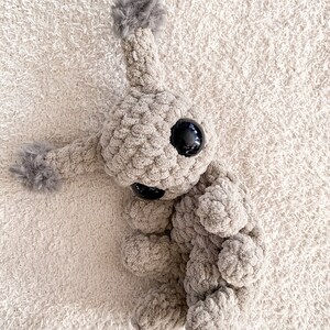 May include: A beige crocheted plush toy with a textured, soft yarn finish. It features a large, round head with a single antenna topped with a gray pom-pom, two large black plastic eyes, a small black nose, and two arms with gray pom-poms.
