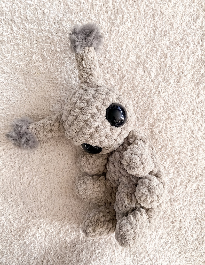May include: A beige crocheted plush toy with a textured, soft yarn finish. It features a large, round head with a single antenna topped with a gray pom-pom, two large black plastic eyes, a small black nose, and two arms with gray pom-poms.