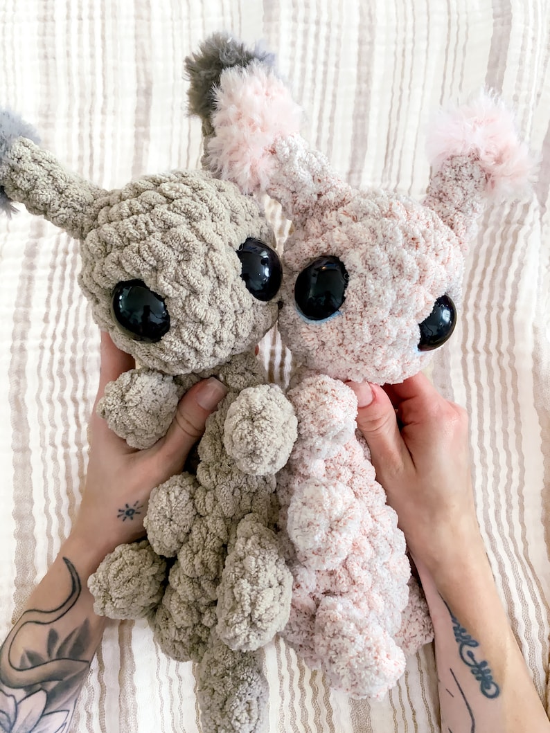 May include: Two plush stuffed animals made of soft, chunky yarn. One is light brown and the other is pale pink. Both feature large, round, glossy black eyes and tufted, fuzzy ears. The creatures have a textured, knotted appearance.