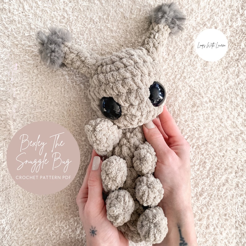 May include: A beige crochet plush toy shaped like a bug with large black plastic eyes, two antennae topped with gray pom-poms, and multiple rounded segments. Text on the image reads Bexley The Snuggle Bug CROCHET PATTERN PDF.