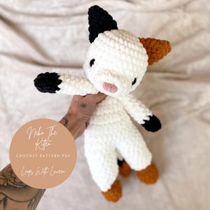 Peut inclure: Un chat en crochet blanc avec des accents noirs et orange. Le chat s'appelle "Neko the Kitten" et le modèle de crochet est disponible à l'achat auprès de "Loops With Lauren".