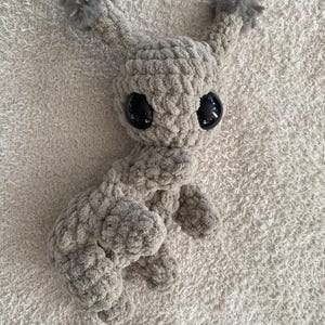 May include: A beige crochet plush toy featuring a rounded head, two tall ears topped with fluffy gray pom-poms, and large, smooth, black plastic safety eyes. The body is crafted from thick, textured yarn with visible stitches.