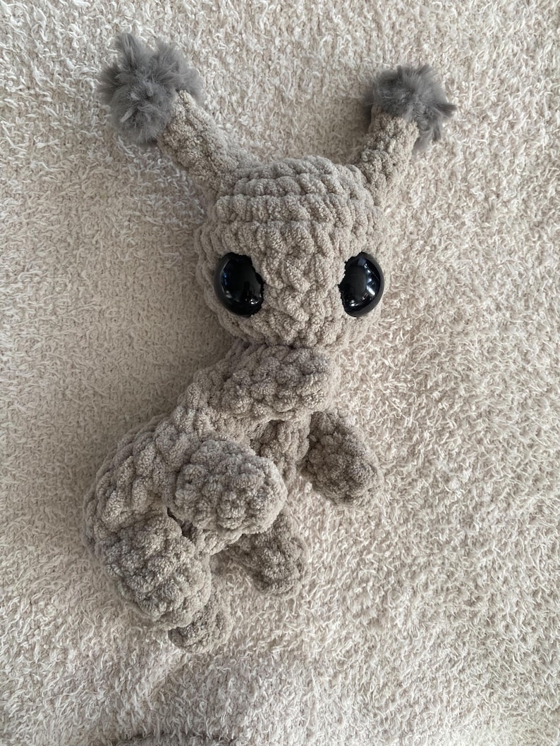 May include: A beige crochet plush toy featuring a rounded head, two tall ears topped with fluffy gray pom-poms, and large, smooth, black plastic safety eyes. The body is crafted from thick, textured yarn with visible stitches.