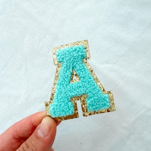May include: A teal chenille letter "A" patch with a gold glitter border. The patch is held up against a white background. The letter is a puffy, textured design.