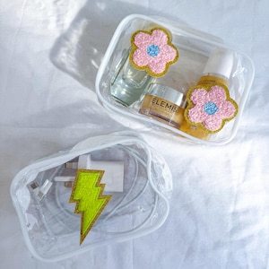 May include: Two clear, rectangular cosmetic bags. One contains skincare products and is decorated with two pink flower patches. The other holds a charger and cord, adorned with a yellow lightning bolt patch. Both bags are on a white surface.