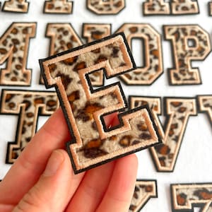 May include: An embroidered letter "E" patch with a leopard print design. The patch has a black border and a tan-coloured outline. Other letter patches are visible in the background.