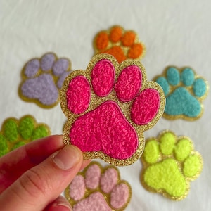 May include: A hand holding a pink paw print with gold glitter edging. Other colourful paw prints in orange, blue, green, purple, and pink are scattered around. These decorative paw prints are likely for crafts or pet-themed projects.