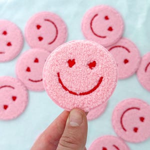 May include: A collection of pink, circular patches, each embroidered with a red smiley face. The patches have a textured, fuzzy appearance. The smiley faces have heart-shaped eyes and a curved mouth. Patches are on a light blue surface.