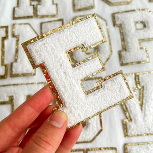 May include: White felt letter "E" with gold glitter edging. The letter is held in a hand, with other white and gold letters in the background. These letters are likely for a jacket or other personalised item.