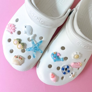 May include: Two white Crocs sandals adorned with decorative charms. Charms include a pink seashell, a white polar bear, a turtle, a blue starfish, a blue whale, a flower, a pink "Summer" sign, a life preserver, a seashell, and a starfish.