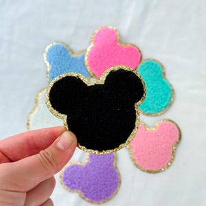 May include: A hand holding a black felt Mickey Mouse head patch with a gold glitter border. Other colourful felt patches in blue, pink, green, purple, and white are scattered in the background.