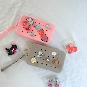 May include: Two rectangular pouches, one pink and one taupe, are decorated with charms. The pink pouch has a strawberry, a chili pepper, and a smiley face charm. The taupe pouch features a white flower, a star, and a dog charm.