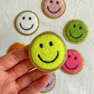 May include: A selection of smiley face patches in various colours. The patches are round and feature a black smiley face on a base of yellow, pink, green, white, and orange. Each patch has a gold glitter border.
