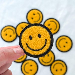 May include: A selection of yellow smiley face patches with black borders and embroidered details. One patch is held by a hand, displaying the textured, fuzzy surface. The patches are arranged on a white background.