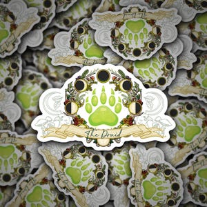 May include: A collection of stickers featuring a central design of a green paw print surrounded by a wreath of moon phases, flowers, and mushrooms. The text "The Druid" is on a banner below the paw print. The stickers have a white border.