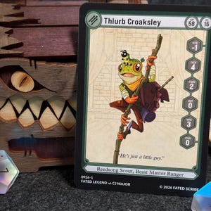 May include: A fantasy card featuring a green frog character named Thlurb Croaksley, holding a staff and wearing a backpack. The card has stats and the text "He's just a little guy." A wooden box with a carved monster face is in the background.