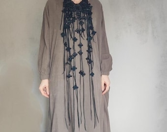 Puede incluir: Un collar largo de cuero negro con un diseño floral. El collar está hecho de múltiples hebras de cuero que están atadas juntas con pequeñas cuentas negras. El collar lo lleva una persona que lleva un vestido marrón.