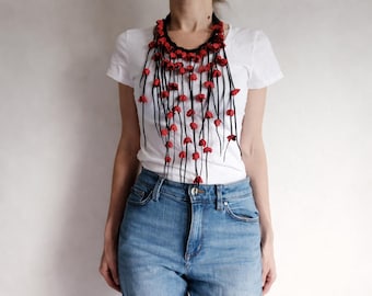 Puede incluir: Un collar hecho a mano con una base negra y hebras colgantes. Cada hebra presenta pequeñas cuentas rojas en forma de flor. El collar se usa sobre una camiseta blanca y unos vaqueros azules.
