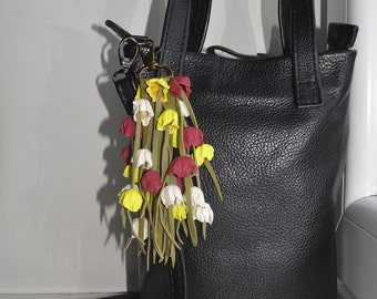 Puede incluir: Un bolso de mano de cuero negro con una correa de hombro desmontable y un charm con forma de flor. El charm está hecho de cuero y tiene flores rojas, amarillas y blancas.