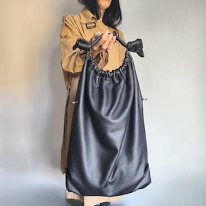Könnte beinhalten: Eine schwarze Leder-Beuteltasche mit geknoteten Griffen. Die Tasche wird von einer Person gehalten, die einen beigen Trenchcoat trägt. Die Tasche hat einen gerafften Oberteil und ein schlichtes, minimalistisches Design, das für den täglichen Gebrauch geeignet ist.