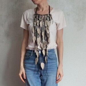 May include: A statement necklace featuring beige leather leaf pendants suspended from black cords. The cords are attached to a dark brown leather collar. The necklace is worn over a light-coloured t-shirt and blue jeans.