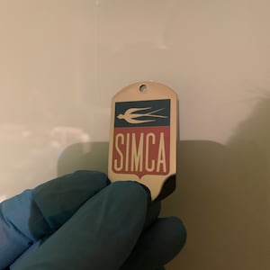 Può includere: Un portachiavi in metallo argentato con un logo blu e rosso che dice "SIMCA" e un design di uccello stilizzato.