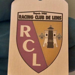 Puede incluir: Un llavero de metal plateado con un emblema marrón y dorado. El emblema presenta el texto "Racing Club de Lens" y las letras "RCL" en blanco. El emblema también presenta un faro y dos flores de lis.