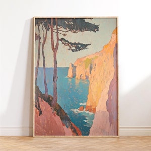 以下が含まれることがあります： 崖、木、海を描いた風景画の額装。青、ピンク、オレンジの色使いで、明るい木製のフレームを使用。絵は白い壁に立てかけられ、木の床に置かれている。