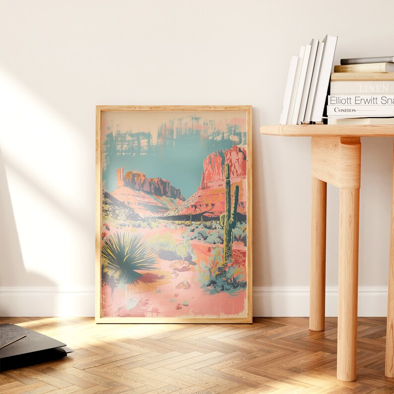 May include: A framed print of a desert landscape with red rock formations, a blue sky, and a tall cactus in the foreground. The print is in a light wood frame.