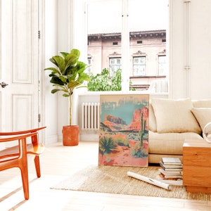 May include: A framed print depicting a desert landscape with red rock formations, cacti, and a blue sky. The print is hanging on a white wall in a living room setting.