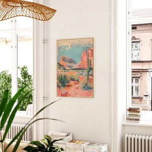 May include: A vintage-style poster depicting a desert landscape with red rock formations, a blue sky, and a cactus in the foreground. The poster has a faded, distressed look and is framed in a light wood frame.