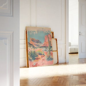 May include: A framed print of a desert landscape with a cactus in the foreground and red rock formations in the background. The print is in a gold frame and is leaning against a white wall.