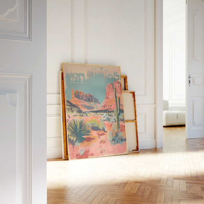 May include: A framed print of a desert landscape with a cactus in the foreground and red rock formations in the background. The print is in a gold frame and is leaning against a white wall.