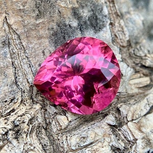 May include: A faceted, pear-shaped gemstone in a vibrant shade of pink. The gem's surface reflects light, showcasing its intricate cuts and internal structure. The gemstone is set against a textured, natural wood background.