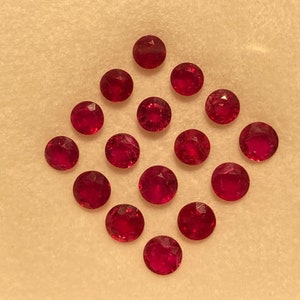Può includere: Una collezione di quattordici gemme rotonde, sfaccettate, rosso intenso, disposte a forma di diamante. Le gemme sono uniformemente distanziate e sembrano lucide, riflettendo la luce. Lo sfondo è di colore beige chiaro.
