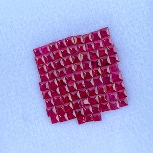 Może przedstawiać: Kolekcja kwadratowo oszlifowanych, ciemnoczerwonych kamieni szlachetnych ułożonych w prostokątny wzór. Kamienie osadzone są na białym tle, tworząc uderzający kontrast. Kamienie szlachetne to prawdopodobnie rubiny, znane z żywego koloru i blasku.