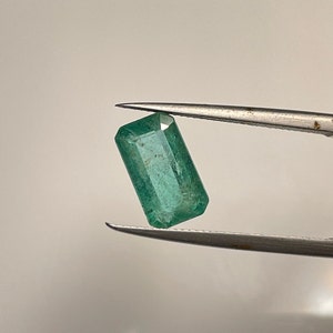 May include: A rectangular-cut, vibrant green emerald gemstone is held by silver tweezers. The gemstone has a slightly translucent appearance, with visible internal inclusions. The emerald is a rich, saturated green colour.