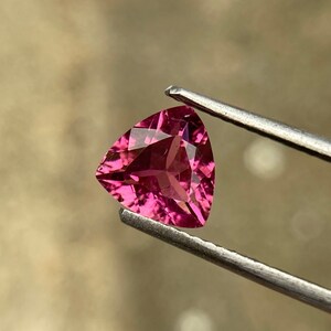 May include: A triangular, pink gemstone is held by tweezers. The gemstone is a vibrant pink colour and has multiple facets that reflect light. The background is a soft, neutral tone. The gemstone is likely a precious stone.