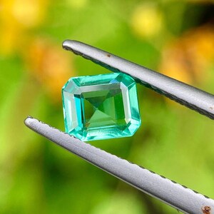 May include: A vibrant, emerald-cut gemstone held by silver tweezers. The gem is a vivid green color with a rectangular shape and faceted edges. The background is a soft, blurred green.