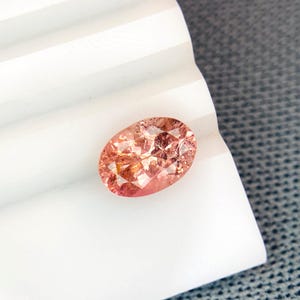 Puede incluir: Una gema ovalada y facetada con una paleta de colores melocotón y rosa. La gema se encuentra sobre una superficie blanca y estriada, con un fondo gris texturizado. Las facetas de la gema capturan la luz, creando un efecto brillante.
