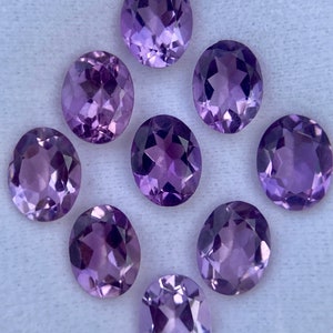 May include: Nine oval-cut amethyst gemstones. The gemstones are a deep purple color and have a faceted surface that reflects light. The stones are arranged in a circular pattern, with one in the center and the others surrounding it.