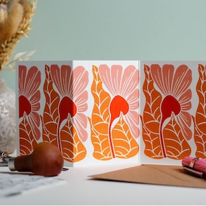 May include: Greeting cards featuring a floral design in pink, orange, and white. The cards are open, showcasing a repeating pattern of large flowers and leaves. A brown envelope and a pink pen are in the foreground.
