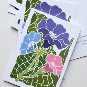 May include: A stack of white greeting cards with a floral design. The cards show a linocut print of morning glory flowers in blue, purple, and pink, with green leaves and stems. The design is repeated on each card.