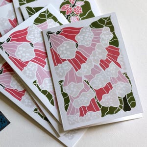 May include: A stack of greeting cards with a floral design. The cards feature a repeating pattern of pink, red, and white flowers with green leaves on a white background. A dark teal blue card with a floral pattern is in the lower left corner.