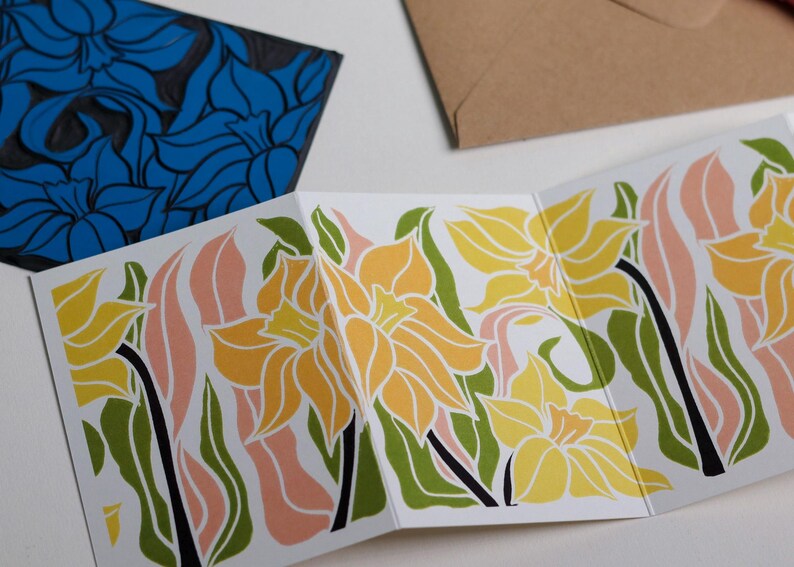 Hand-carved lino block shown beside finished Daffodil Garland card. Traditional linocut printmaking tools and original carved linoleum block displayed with a contemporary botanical portrait card. No AI, authentic craftsmanship from Norfolk studio