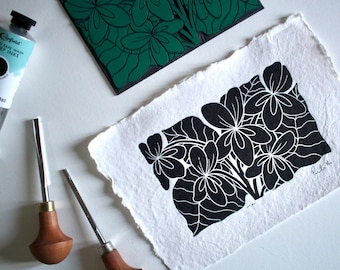 May include: Black and white linocut print of a cluster of flowers with leaves. The print is on white paper and has a textured surface. The artist's signature is in the bottom right corner.