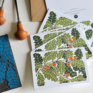 May include: A collection of greeting cards featuring a green and orange oak leaf design. The cards are stacked on a white surface, alongside a blue linocut block and carving tools with wooden handles. The design includes acorns.